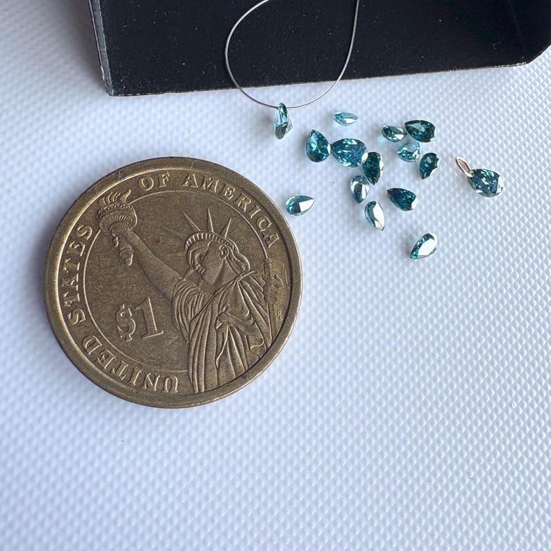 May include: A collection of small, teal-colored gemstones in a teardrop shape, scattered on a white surface. A US one-dollar coin is next to the gemstones for size comparison. A black box is in the upper left corner.