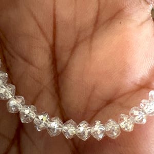 Puede incluir: Un brazalete de cuentas transparentes y facetadas. Las cuentas en forma de diamante est&aacute;n ensartadas. El brazalete se sostiene en una mano, con el tono de la piel como fondo c&aacute;lido. Las cuentas reflejan la luz, creando un efecto brillante.