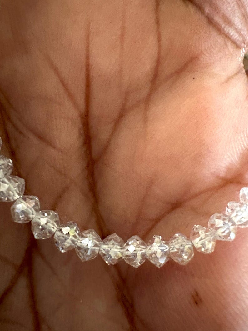 Puede incluir: Un brazalete de cuentas transparentes y facetadas. Las cuentas en forma de diamante est&aacute;n ensartadas. El brazalete se sostiene en una mano, con el tono de la piel como fondo c&aacute;lido. Las cuentas reflejan la luz, creando un efecto brillante.
