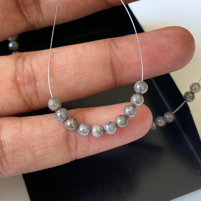 May include: A strand of small, round, gray beads on a thin, silver wire. The beads have a textured surface and are arranged in a curved line. The background is a dark, blurred surface.