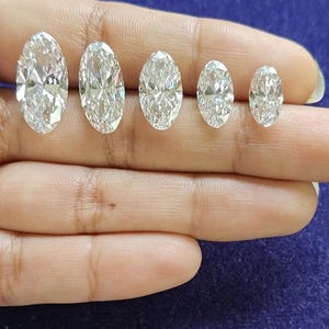 May include: Five clear, colorless, faceted gemstones with an oval cut, arranged in a horizontal row from largest to smallest. Each stone features a brilliant, reflective surface with multiple facets that catch and refract light, creating sparkle.