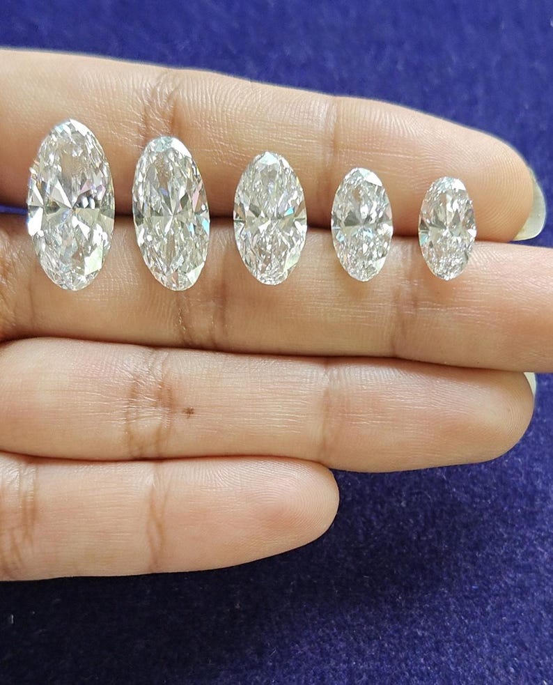 May include: Five clear, colorless, faceted gemstones with an oval cut, arranged in a horizontal row from largest to smallest. Each stone features a brilliant, reflective surface with multiple facets that catch and refract light, creating sparkle.