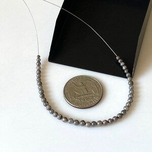 May include: A strand of silver-grey beads strung on a thin wire. The beads are small and round, and they are arranged in a circle. A US quarter is included for scale.