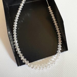 Puede incluir: Un delicado collar compuesto por cuentas claras y facetadas ensartadas en un fino cord&oacute;n blanco. El collar se exhibe dentro de una caja negra, con las cuentas captando la luz y creando un efecto brillante. El dise&ntilde;o general es simple y elegante.