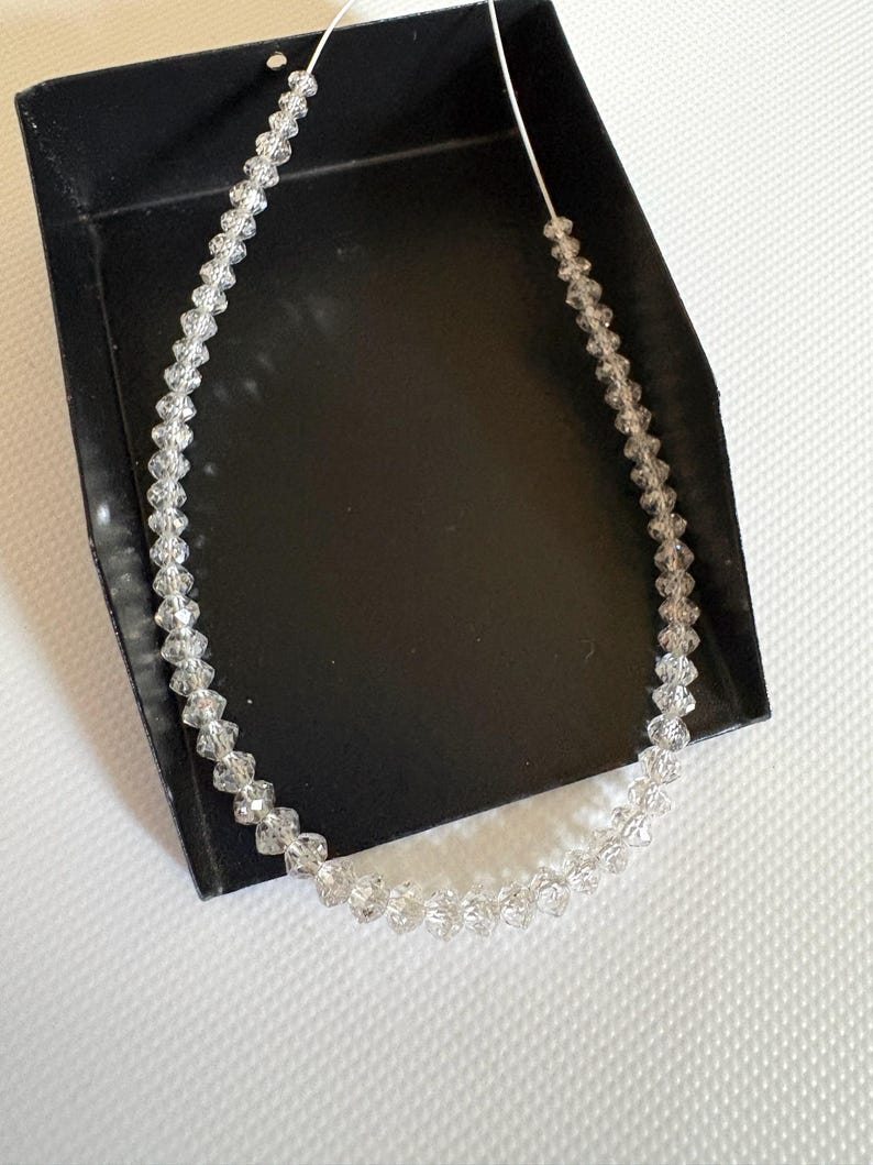 Puede incluir: Un delicado collar compuesto por cuentas claras y facetadas ensartadas en un fino cord&oacute;n blanco. El collar se exhibe dentro de una caja negra, con las cuentas captando la luz y creando un efecto brillante. El dise&ntilde;o general es simple y elegante.