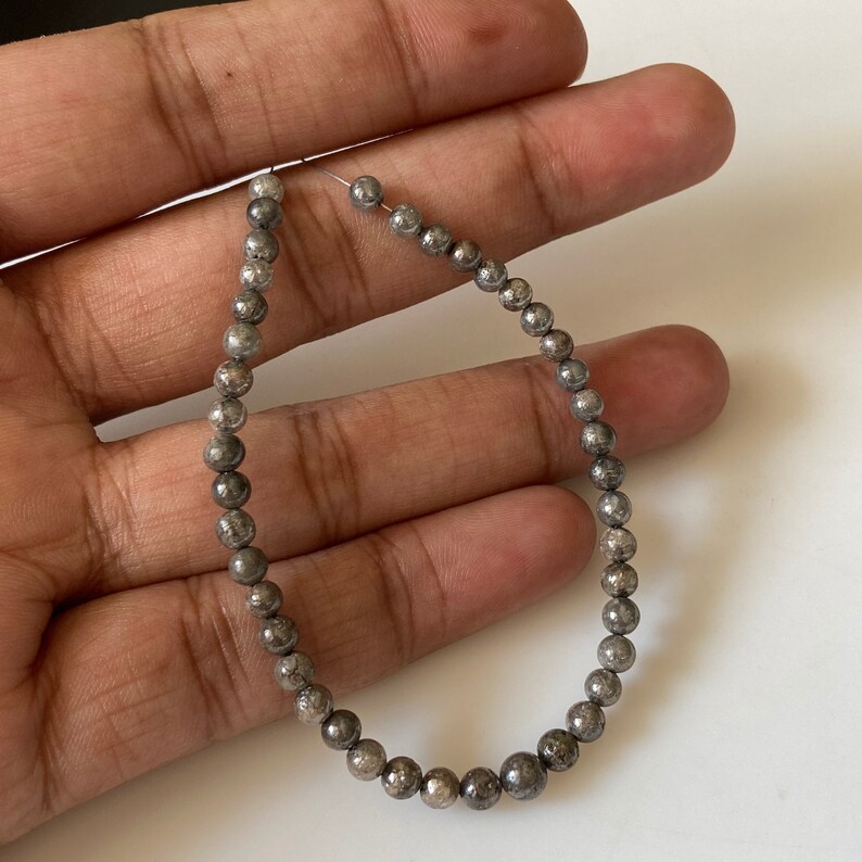 May include: Strand of small, round, metallic gray beads. The beads are strung on a thin, clear wire, forming a loop. The beads have a slightly textured surface and a subtle sheen. The background is a neutral, off-white color.