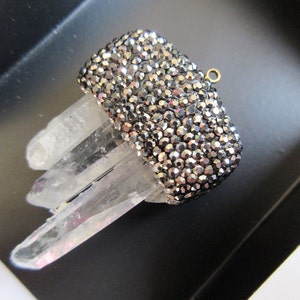 May include: A rectangular, silver-toned, crystal-covered object with a small gold loop, resting on clear quartz crystals. The crystals are set against a black background, creating a contrast of textures and colors.