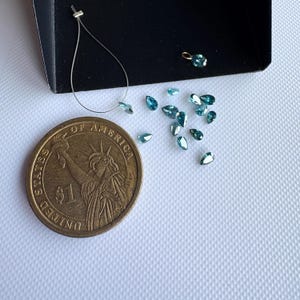 May include: A collection of teal-colored, pear-shaped gemstones scattered on a white surface, next to a US one-dollar coin. A small black tray holds more gems and a partially visible necklace.