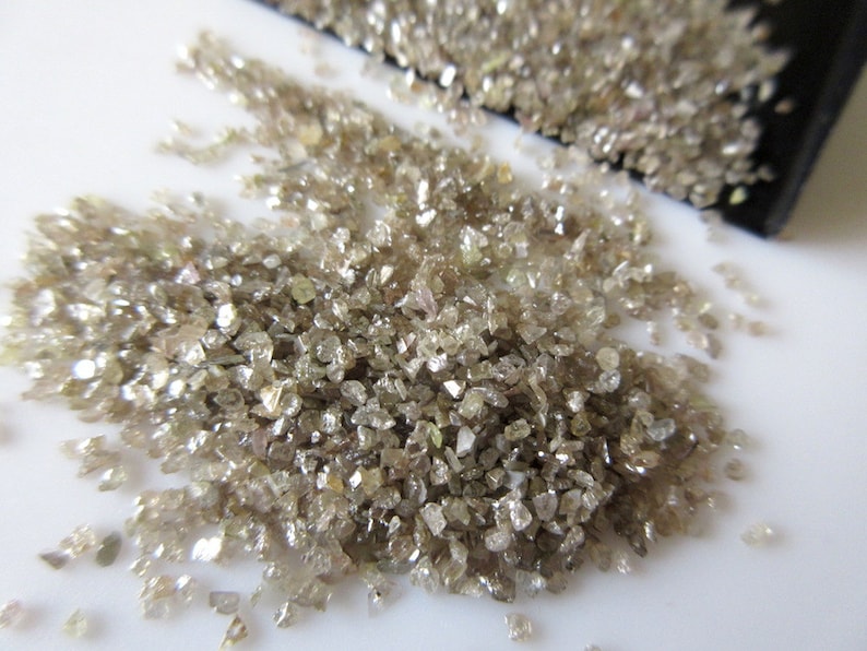 May include: A pile of small, rough, light brown diamonds. The diamonds are scattered on a white surface.