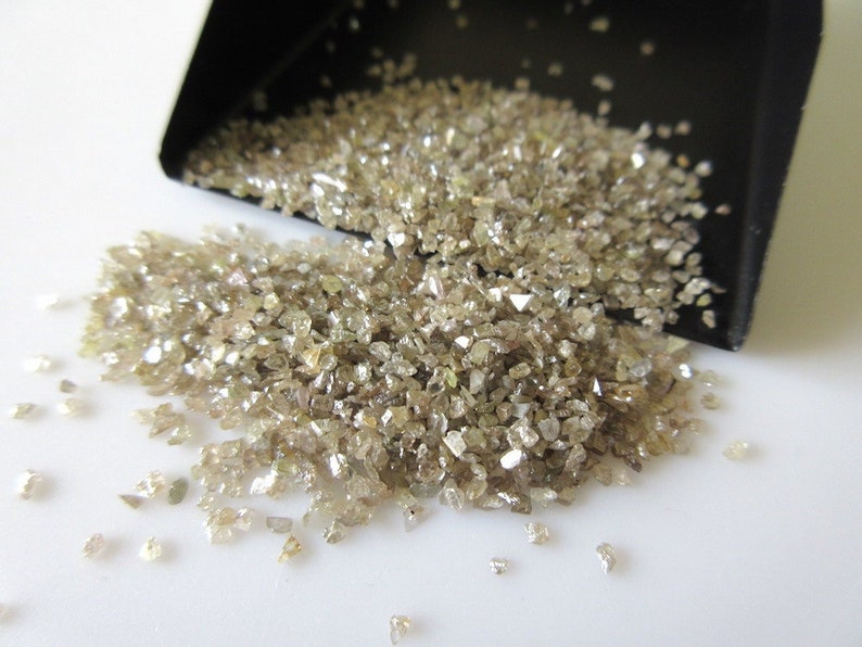 May include: A small scoop filled with a pile of light brown, rough, uncut diamonds. The diamonds are scattered on a white surface.
