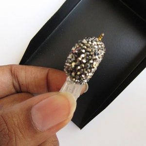 May include: A small, decorative object with a rounded, crystal-covered top and a clear, rectangular base. The top is covered in small, sparkling crystals in shades of silver and gray. A small gold-colored metal piece is at the top. The object is held in a black tray.