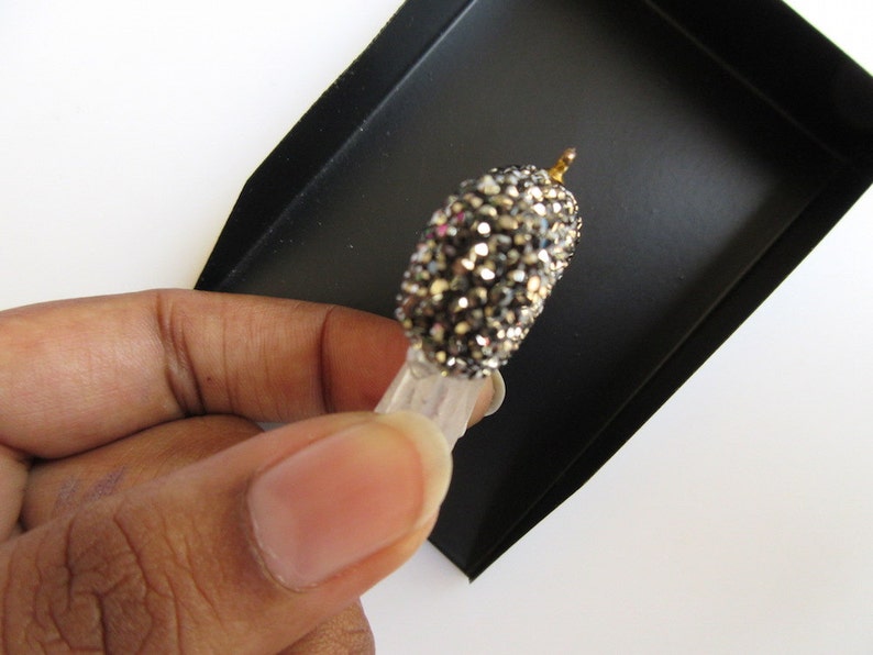 May include: A small, decorative object with a rounded, crystal-covered top and a clear, rectangular base. The top is covered in small, sparkling crystals in shades of silver and gray. A small gold-colored metal piece is at the top. The object is held in a black tray.