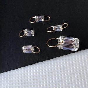 May include: Five clear, rectangular-cut gemstones with gold-colored loops. The gemstones are arranged on a dark surface. The largest gemstone is in the foreground, with the others scattered around it. The gemstones are faceted, reflecting light.