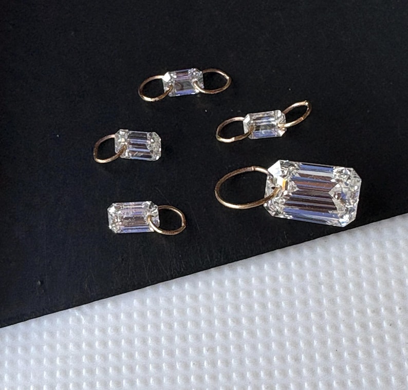 May include: Five clear, rectangular-cut gemstones with gold-colored loops. The gemstones are arranged on a dark surface. The largest gemstone is in the foreground, with the others scattered around it. The gemstones are faceted, reflecting light.