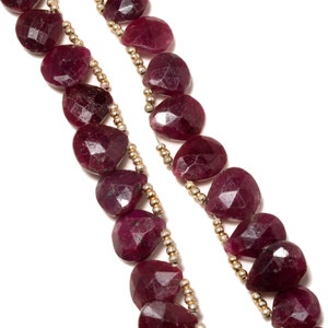 Pode incluir: Um fio de contas de pedras preciosas de cor vermelha rubi com acabamento facetado. As contas são enfiadas em um fio de prata com pequenas contas douradas entre cada pedra preciosa.