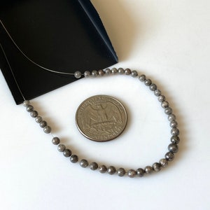 May include: Strung, small, round, gray beads are displayed next to a U.S. quarter coin. The beads are on a thin, silver wire. The beads are a metallic gray color. The background is white with a black tray in the upper left corner.