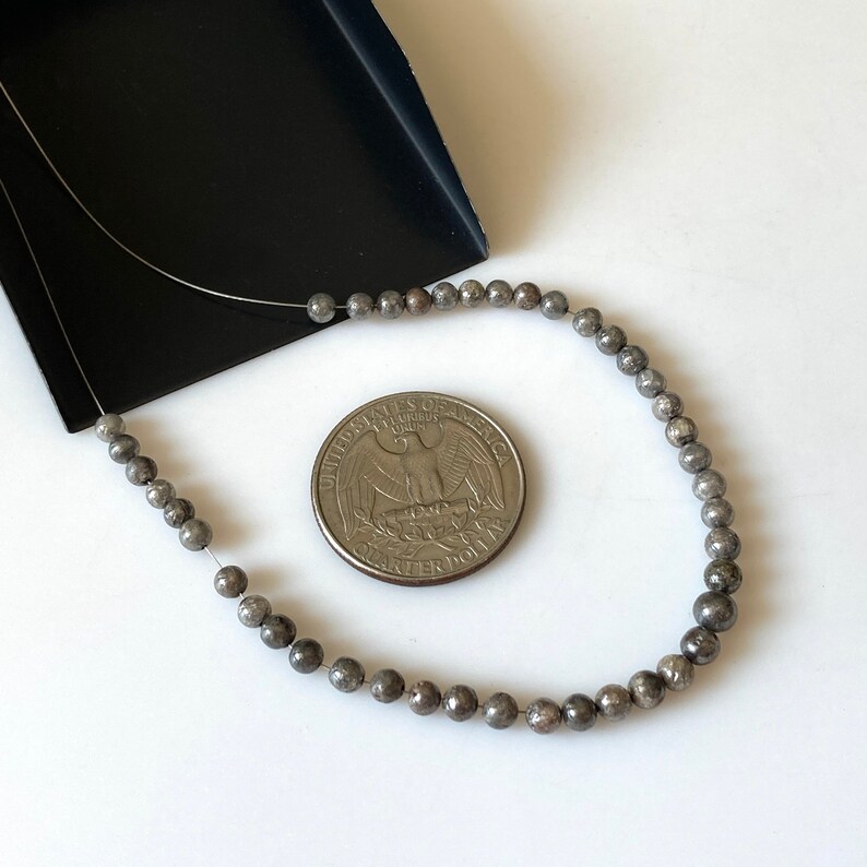 May include: Strung, small, round, gray beads are displayed next to a U.S. quarter coin. The beads are on a thin, silver wire. The beads are a metallic gray color. The background is white with a black tray in the upper left corner.