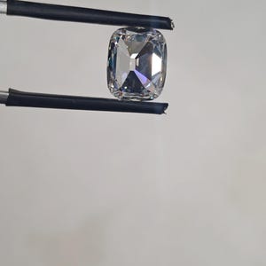 May include: A large, clear, cushion-cut gemstone held by black tweezers. The gemstone has a square shape with rounded corners and multiple facets that reflect light. The background is a plain, light grey.