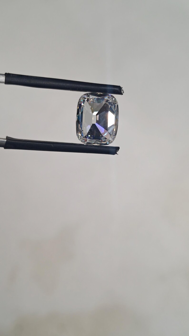 May include: A large, clear, cushion-cut gemstone held by black tweezers. The gemstone has a square shape with rounded corners and multiple facets that reflect light. The background is a plain, light grey.