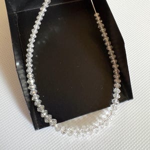 Puede incluir: Un collar de cristal transparente con cuentas facetadas ensartadas en un cord&oacute;n fino y transparente. El collar se exhibe en una caja negra sobre un fondo blanco. Las cuentas est&aacute;n espaciadas uniformemente, creando un efecto brillante.