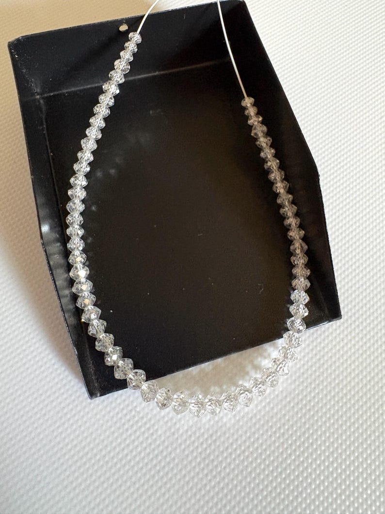 Puede incluir: Un collar de cristal transparente con cuentas facetadas ensartadas en un cord&oacute;n fino y transparente. El collar se exhibe en una caja negra sobre un fondo blanco. Las cuentas est&aacute;n espaciadas uniformemente, creando un efecto brillante.
