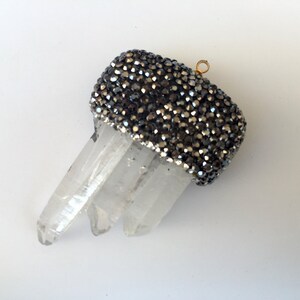 May include: A clear quartz crystal pendant with three points. The top is adorned with a band of small, dark gray, sparkling beads. A small gold-colored loop is attached for hanging.