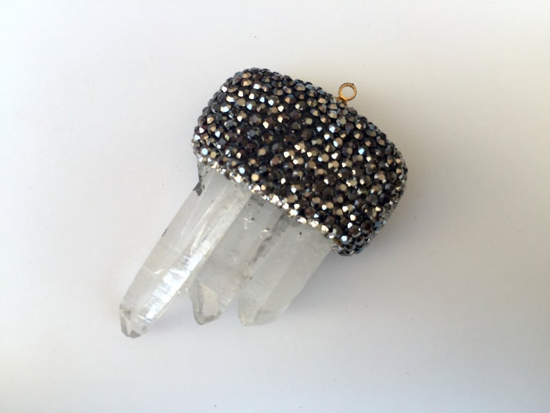 May include: A clear quartz crystal pendant with three points. The top is adorned with a band of small, dark gray, sparkling beads. A small gold-colored loop is attached for hanging.
