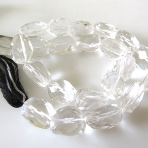 K&ouml;nnte beinhalten: Eine Kette aus klaren Quarzkristallperlen, jede Perle ist facettiert und oval geformt. Die Perlen sind auf einer schwarzen und wei&szlig;en Schnur mit einem Quasten aufgef&auml;delt.