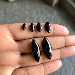 以下が含まれることがあります： 手の上に置かれた、さまざまなサイズの黒い宝石のカボションが5つ。カボションはすべて楕円形で、滑らかで磨かれた仕上げになっています。