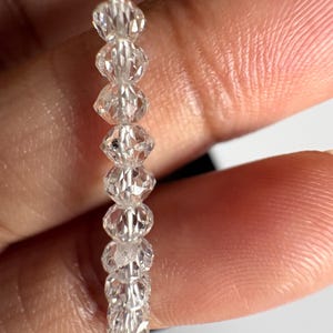 Puede incluir: Un hilo de cuentas de cristal transparente y facetado. Las cuentas est&aacute;n ensartadas en un fino alambre plateado. Los cristales tienen un corte en forma de diamante, que refleja la luz. Las cuentas se sostienen en una mano.