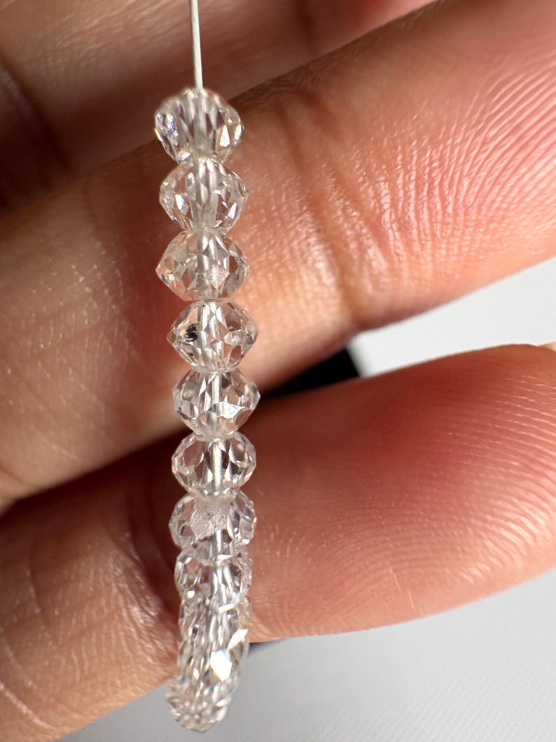 Puede incluir: Un hilo de cuentas de cristal transparente y facetado. Las cuentas est&aacute;n ensartadas en un fino alambre plateado. Los cristales tienen un corte en forma de diamante, que refleja la luz. Las cuentas se sostienen en una mano.
