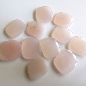 May include: A collection of ten smooth, pale pink, square-shaped stones. The stones are arranged in a cluster on a white surface.