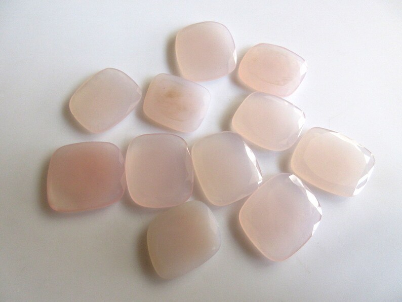 May include: A collection of ten smooth, pale pink, square-shaped stones. The stones are arranged in a cluster on a white surface.