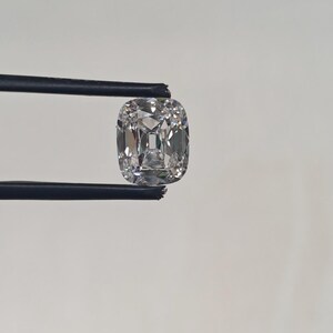 May include: A cushion-cut diamond held by black tweezers. The diamond is clear and faceted, reflecting light. The shape is square with rounded corners, showcasing its brilliance. The background is a plain white.