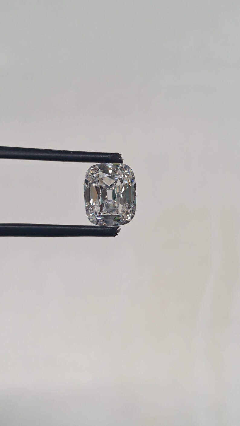 May include: A cushion-cut diamond held by black tweezers. The diamond is clear and faceted, reflecting light. The shape is square with rounded corners, showcasing its brilliance. The background is a plain white.