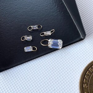 May include: Close-up of several small, rectangular, clear crystal charms with gold-colored loops. The charms are displayed on a dark surface, with a coin visible in the lower right corner. The crystals have a faceted cut.