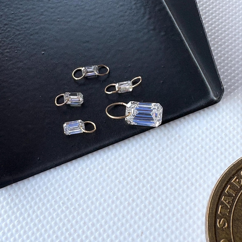 May include: Close-up of several small, rectangular, clear crystal charms with gold-colored loops. The charms are displayed on a dark surface, with a coin visible in the lower right corner. The crystals have a faceted cut.