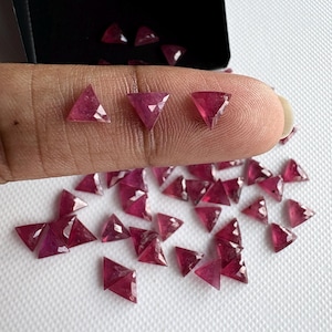 May include: A collection of triangular, faceted, deep red gemstones. The gems are scattered on a white surface, with some held by a finger. The gems have a polished, reflective surface, and are likely rubies.