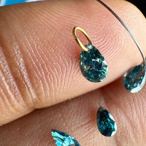 May include: A collection of pear-shaped, deep teal-colored gemstones with faceted surfaces. One gemstone features a small, circular gold-toned metal bail attached to its top, while others are loose and scattered nearby.