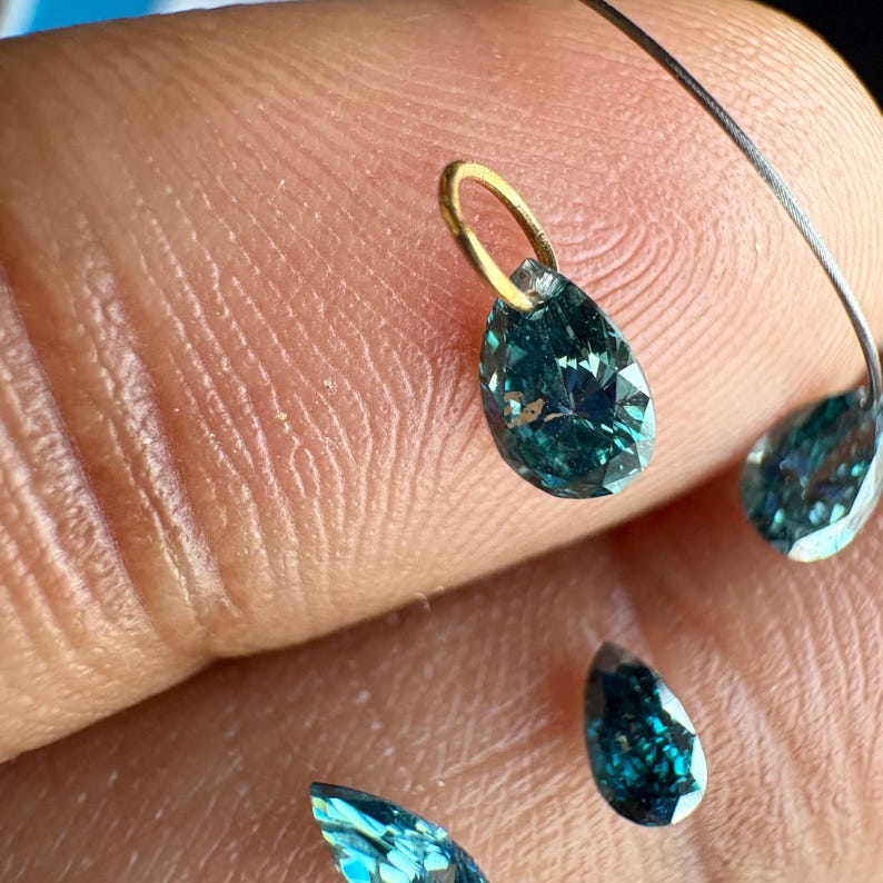May include: A collection of pear-shaped, deep teal-colored gemstones with faceted surfaces. One gemstone features a small, circular gold-toned metal bail attached to its top, while others are loose and scattered nearby.