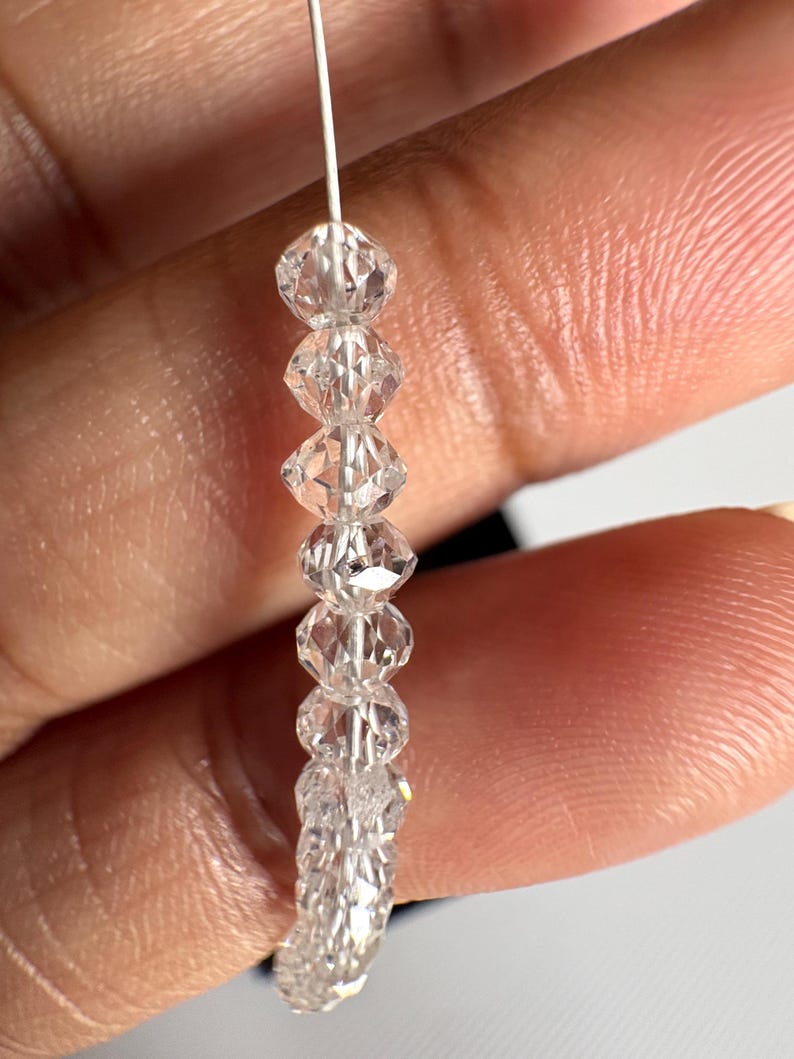 Puede incluir: Un hilo de cuentas de cristal transparente y facetado. Las cuentas est&aacute;n ensartadas en un fino alambre plateado. Los cristales tienen forma de diamante y reflejan la luz, creando un efecto brillante. Las cuentas se sostienen sobre un fondo borroso.