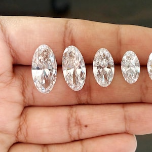 May include: Five clear, colorless, oval-cut gemstones arranged in a row by size from largest to smallest. Each stone features multiple facets that reflect light, creating a brilliant, sparkling appearance against a neutral skin tone background.