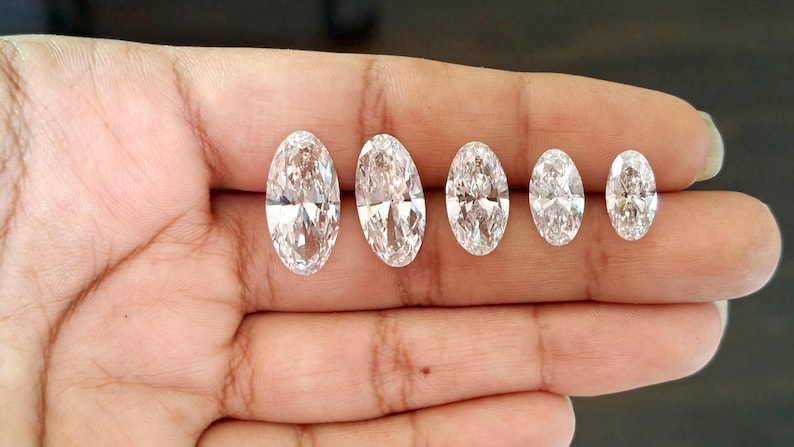 May include: Five clear, colorless, oval-cut gemstones arranged in a row by size from largest to smallest. Each stone features multiple facets that reflect light, creating a brilliant, sparkling appearance against a neutral skin tone background.