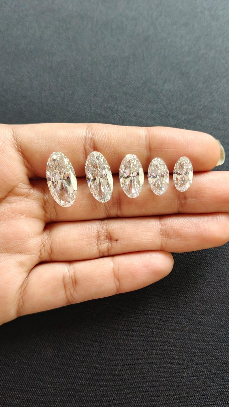 May include: Five clear, oval-cut gemstones arranged in a row by size from largest to smallest. Each stone features a faceted surface that reflects light, creating a brilliant, sparkling appearance against the dark background.