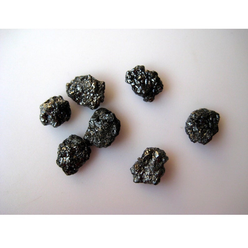 Puede incluir: Una colecci&oacute;n de siete diamantes negros, cada uno con una forma y un tama&ntilde;o &uacute;nicos, presentados sobre una superficie blanca.