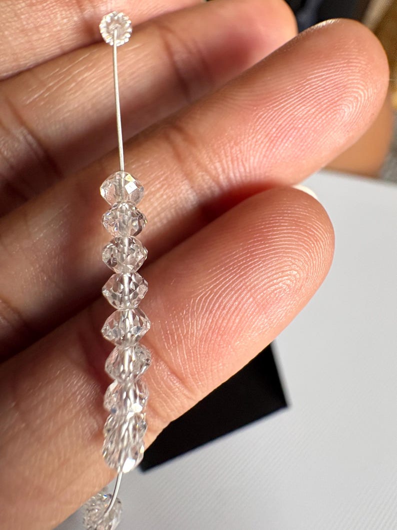 Puede incluir: Una delicada hebra de cuentas de cristal transparente. La hebra presenta m&uacute;ltiples cuentas de cristal facetadas ensartadas en un fino alambre plateado. Las cuentas se mantienen unidas por un peque&ntilde;o cristal redondo en la parte superior. Los cristales brillan.