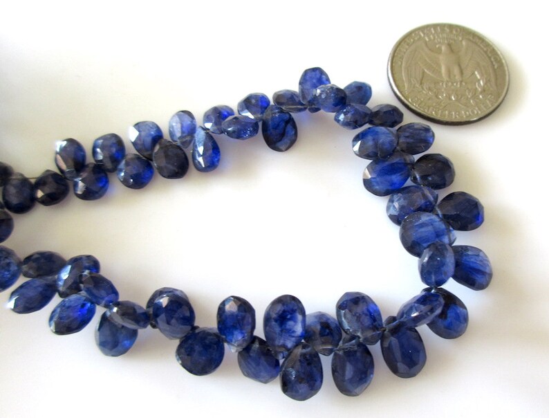 May include: A strand of dark blue gemstone beads, each bead is faceted and oval shaped. The beads are approximately the size of a US quarter.