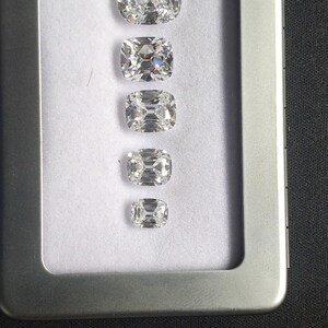 May include: Five clear, cushion-cut gemstones of different sizes are displayed in a row inside a silver metal case. The gemstones sparkle, revealing their facets against a white backdrop. The rectangular case has a hinged lid, showcasing the gems' brilliance.