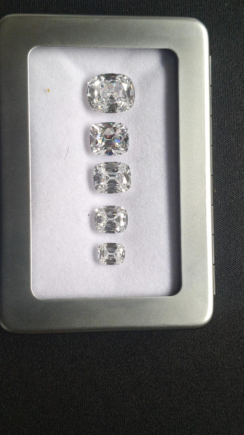 May include: Five clear, cushion-cut gemstones of different sizes are displayed in a row inside a silver metal case. The gemstones sparkle, revealing their facets against a white backdrop. The rectangular case has a hinged lid, showcasing the gems' brilliance.