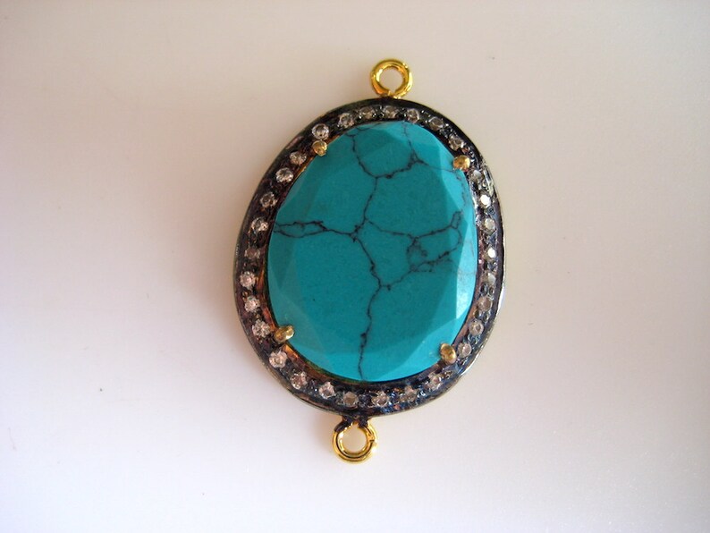 May include: A turquoise gemstone pendant with a silver and gold setting. The pendant is oval shaped and has a detailed design. It is surrounded by small clear stones.